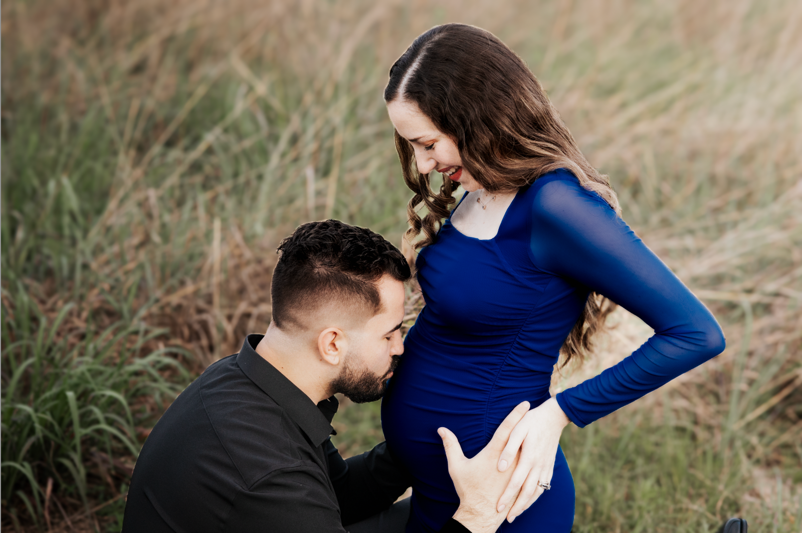 Man kneeling to kiss his pregnant partner's belly in a grassy field