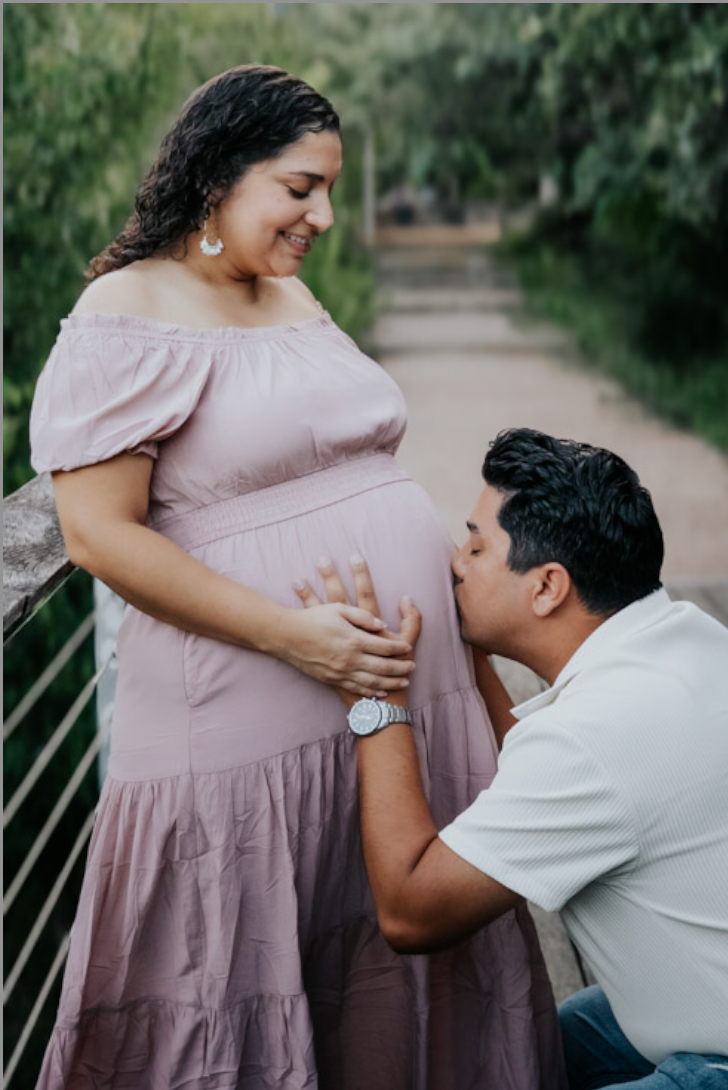 Man kneeling to kiss his partner's pregnant belly on a garden path