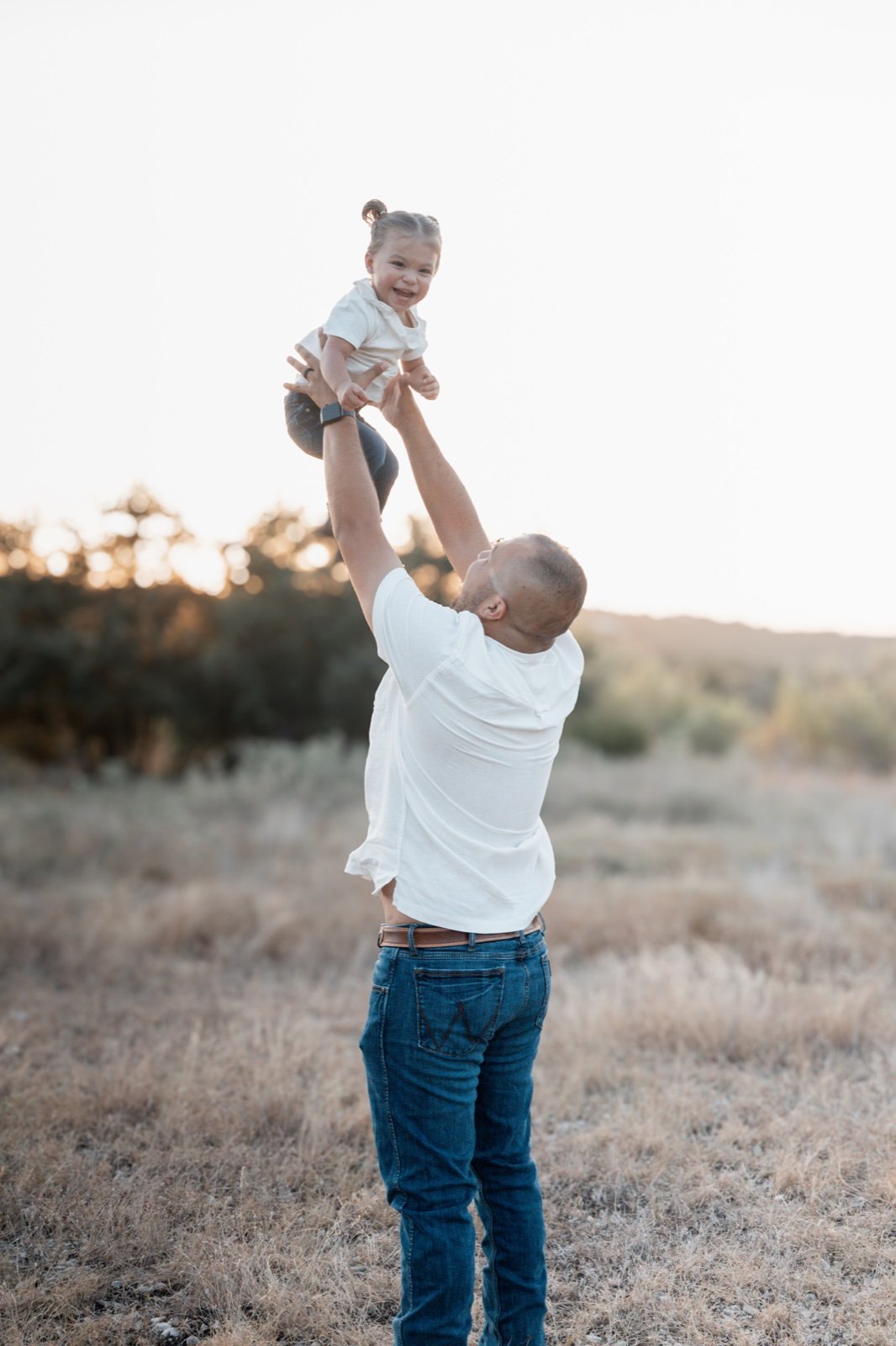 Dad joyfully tossing his laughing toddler in the air in a golden-hour field