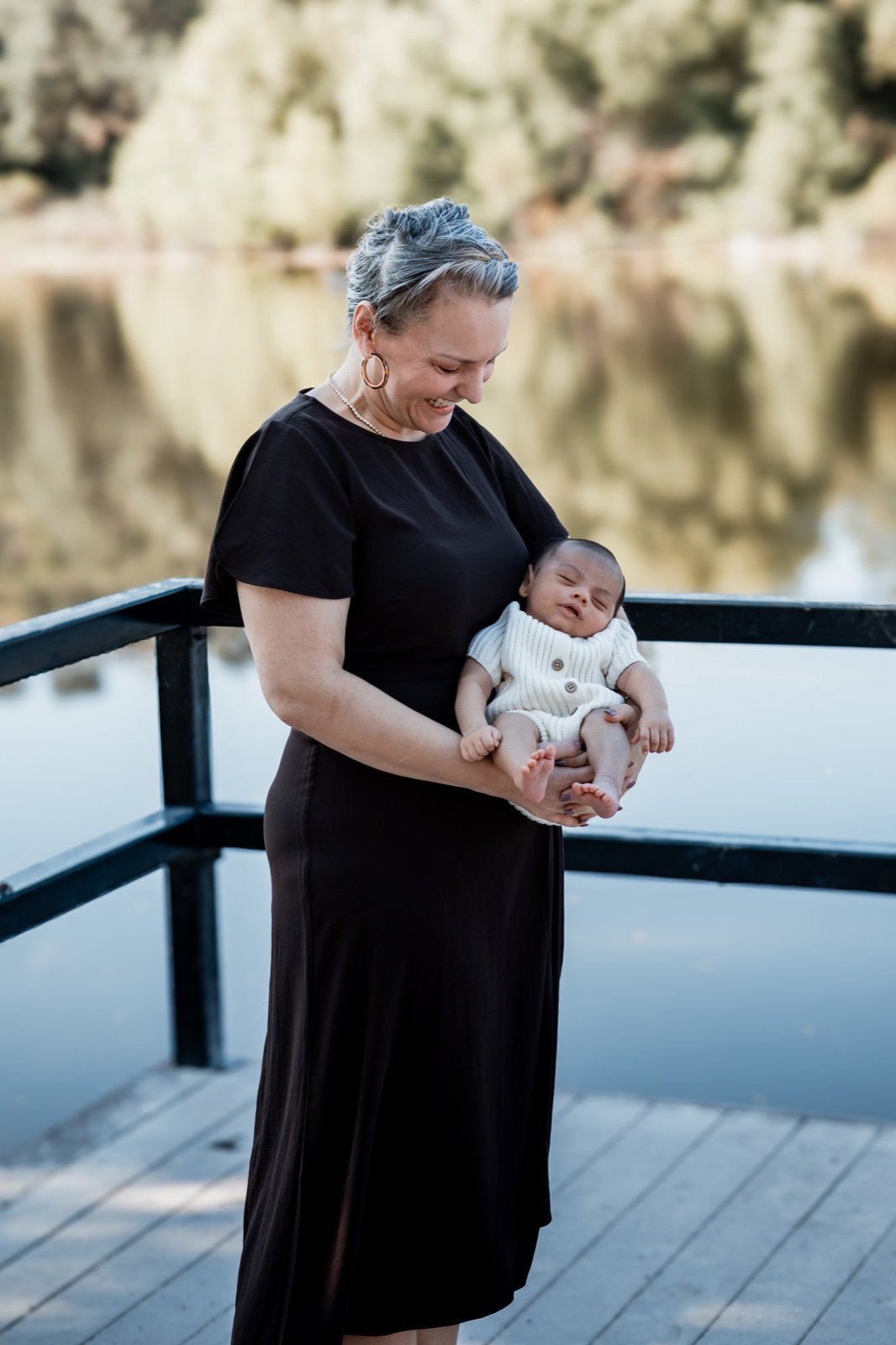 Mother in a black dress cradling her sleeping newborn on a dock by the water